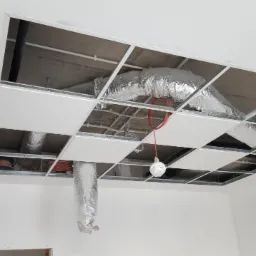 Montaż sufitu podwieszanego kasetonowego z widoczną konstrukcją metalową, instalacją wentylacyjną w folii aluminiowej i czujnikiem dymu, częściowo wypełnionego białymi płytami.