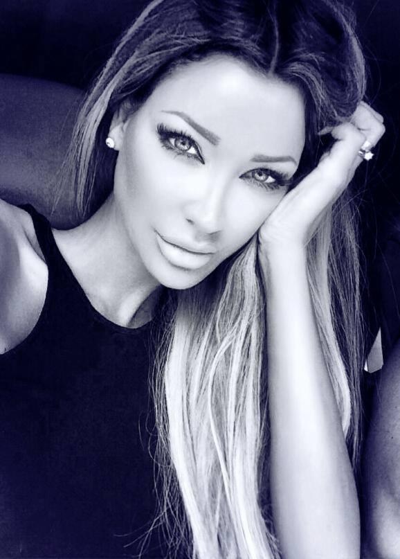 Portret kobiety z długimi, blond włosami, opierającej głowę na dłoni, z wyrazistym makijażem oczu i ust, w czarnej bluzce na tle ciemnego otoczenia.