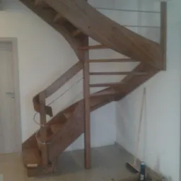 Spiralne schody drewniane z metalową poręczą, w trakcie montażu. Widoczne narzędzia i materiały na podłodze. Ujęcie od dołu, ukazujące konstrukcję schodów.