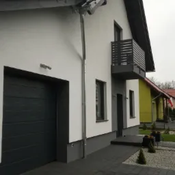 Wykończona elewacja domu jednorodzinnego w kolorze białym z szarą stolarką okienną i bramą garażową, widoczny balkon z czarną balustradą i elementy systemu rynnowego.