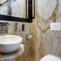 Galeria Kamienia Kamil Lipiński - Biała, okrągła umywalka nablatowa na blacie z beżowego marmuru w łazience z marmurowymi ścianami, widoczne również lustro w czarnej ramie i biały panel spłuczki.