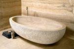 Owalna, kamienna wanna wolnostojąca w beżowym kolorze, z widoczną armaturą i ręcznikiem, na tle ściany wyłożonej płytami z naturalnego kamienia.