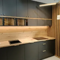 Nowoczesna kuchnia z ciemnoszarymi szafkami, drewnianym blatem i panelem ściennym, oświetleniem LED oraz dekoracyjną ścianką z drewnianych listew, prezentowana w salonie meblowym.