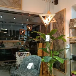 Wnętrze sklepu meblowego w Nowym Targu: krzesła z etykietami cenowymi, sztuczna roślina w plecionym koszu, drewniana lampa wisząca o geometrycznym kształcie i dekoracyjny panel ścienny.