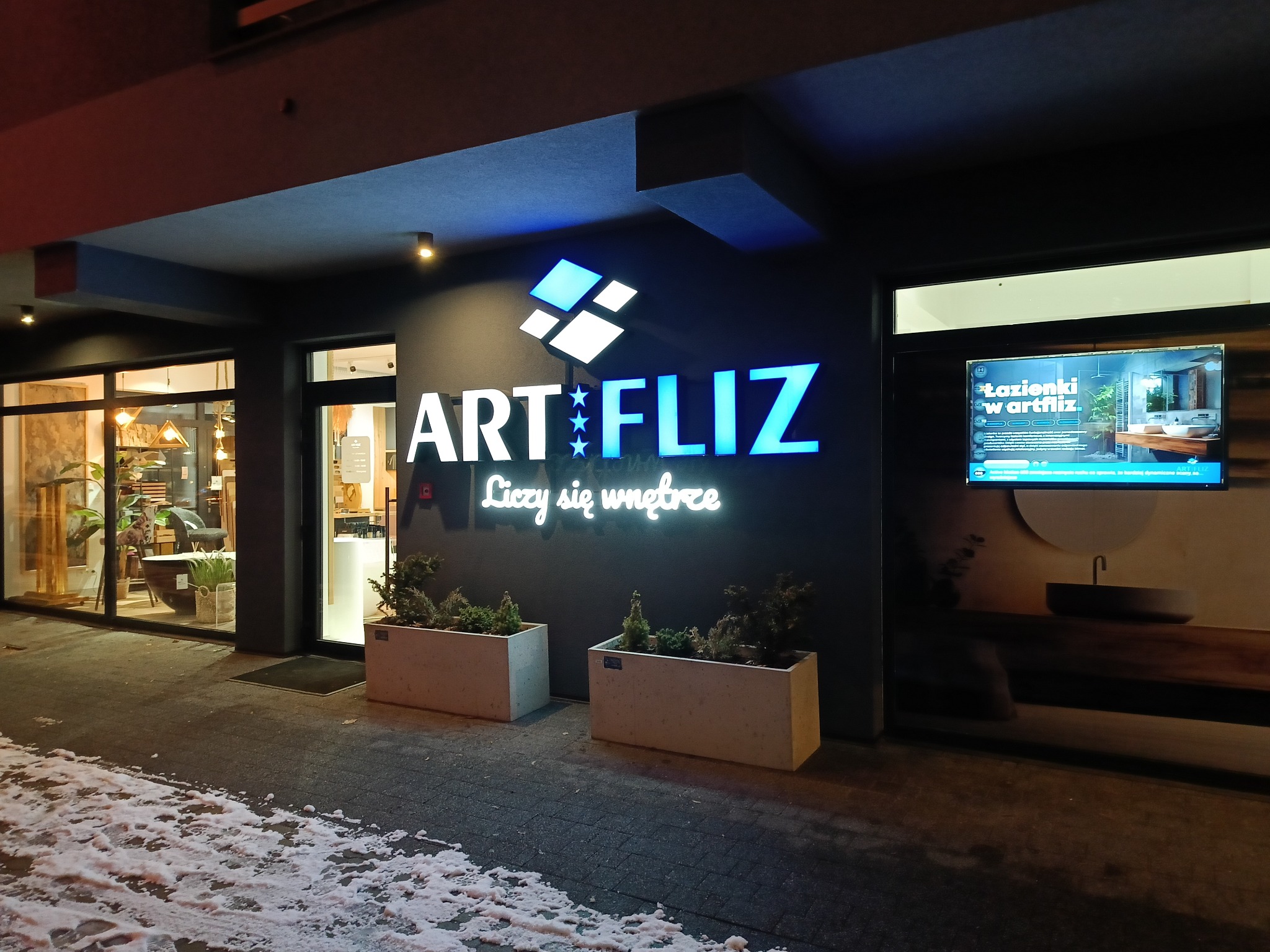 Fasada budynku firmy ArtFliz z podświetlanym logo i napisem 'Liczy się wnętrze', dwa betonowe kwietniki z roślinami przed wejściem, fragment chodnika pokryty śniegiem, w oknach widoczne aranżacje...
