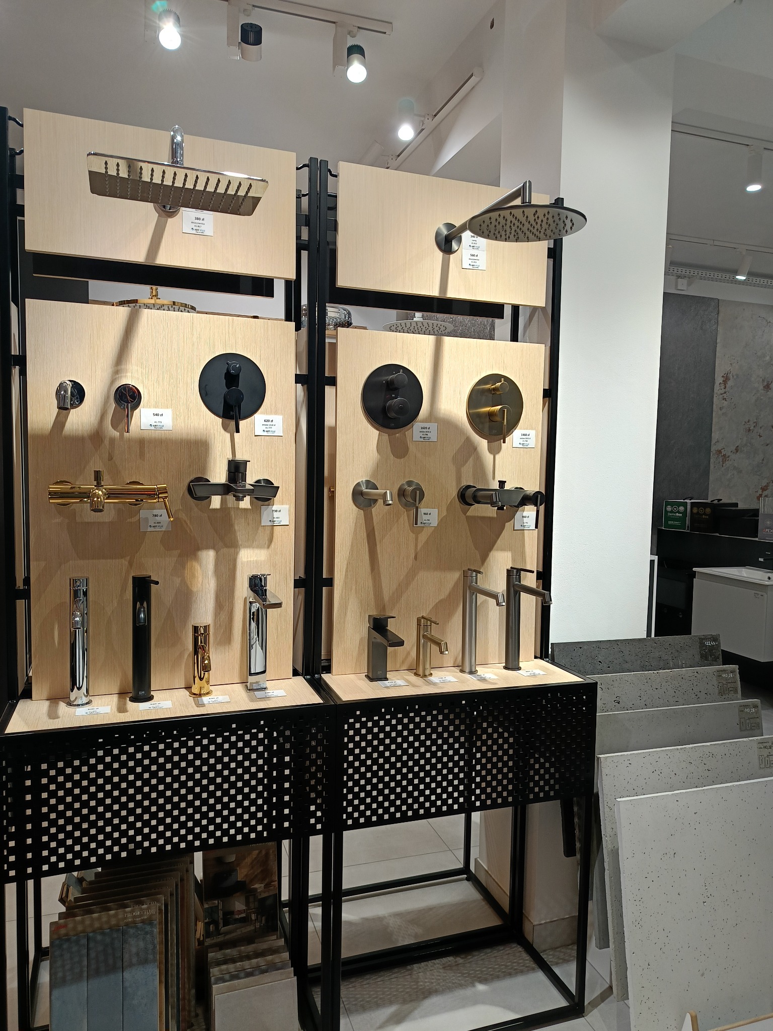 Ekspozycja różnorodnych baterii i głowic prysznicowych w salonie, prezentująca modele w kolorach chromu, czerni i złota na drewnianych panelach z czarnymi metalowymi ramami.