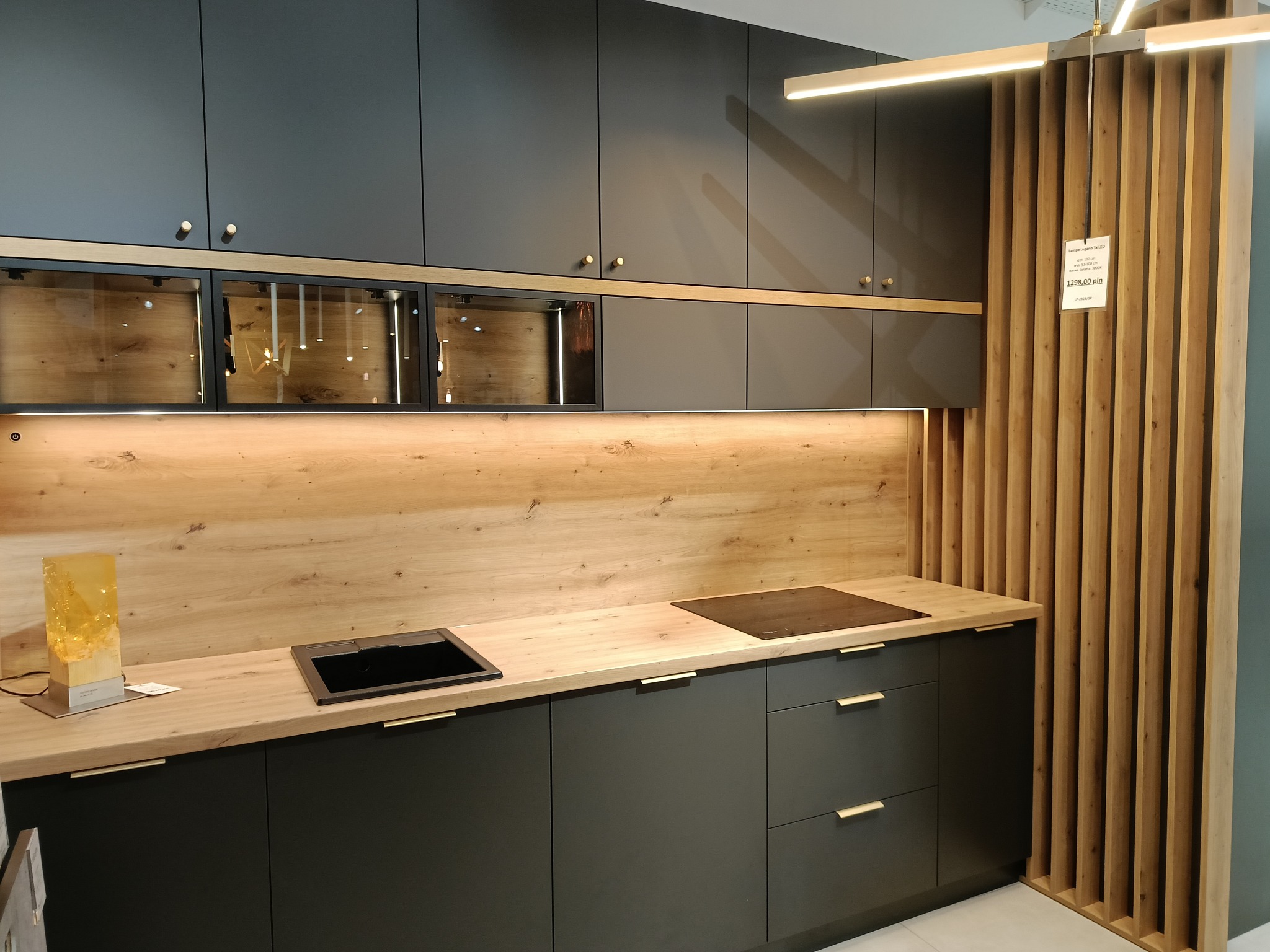 Nowoczesna kuchnia z ciemnoszarymi szafkami, drewnianym blatem i panelem ściennym, oświetleniem LED oraz dekoracyjną ścianką z drewnianych listew, prezentowana w salonie meblowym.