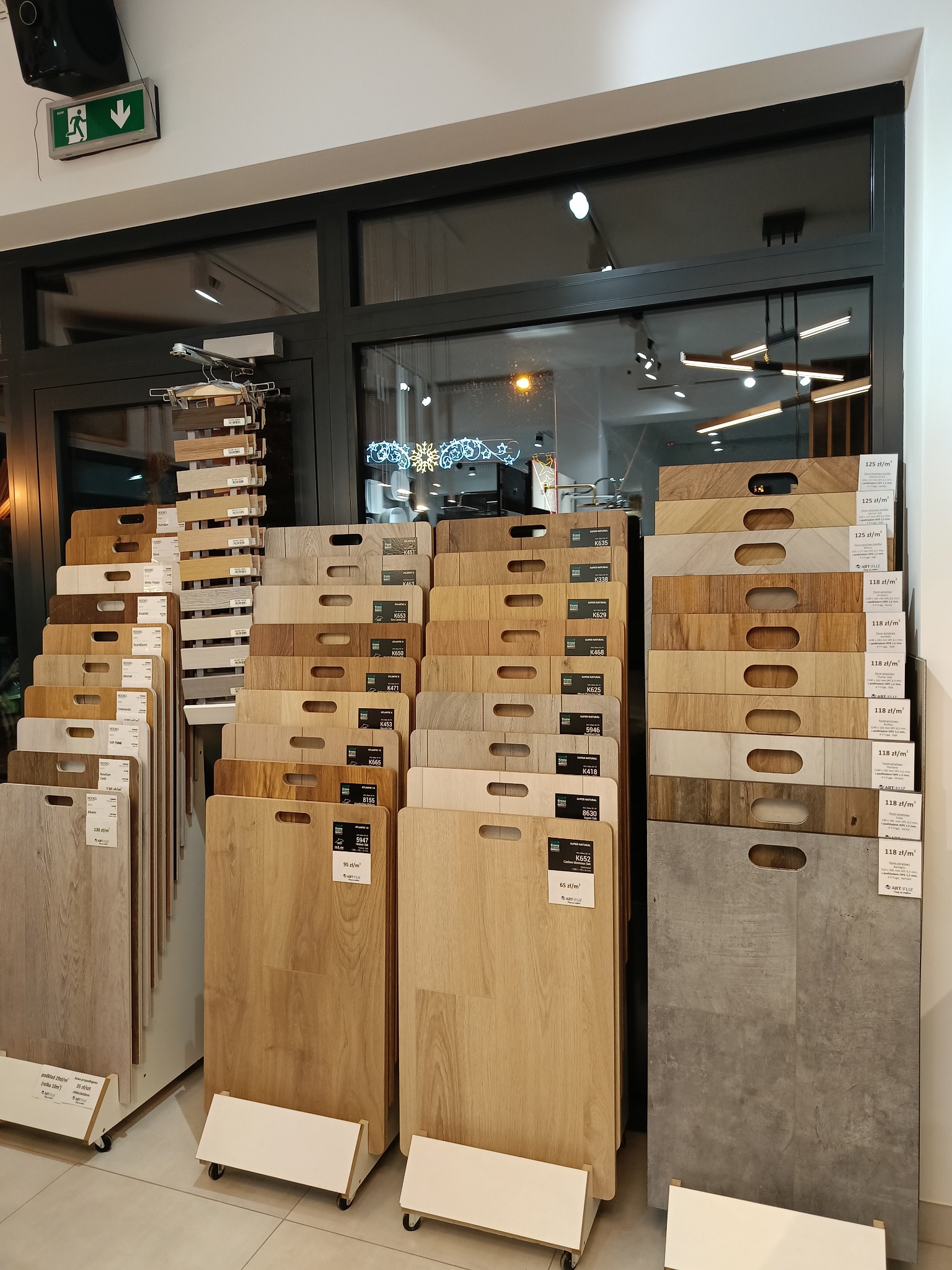 Ekspozycja próbek paneli podłogowych w sklepie, różne odcienie drewna i szarości, widoczne ceny za metr kwadratowy, oświetlenie sufitowe w tle.