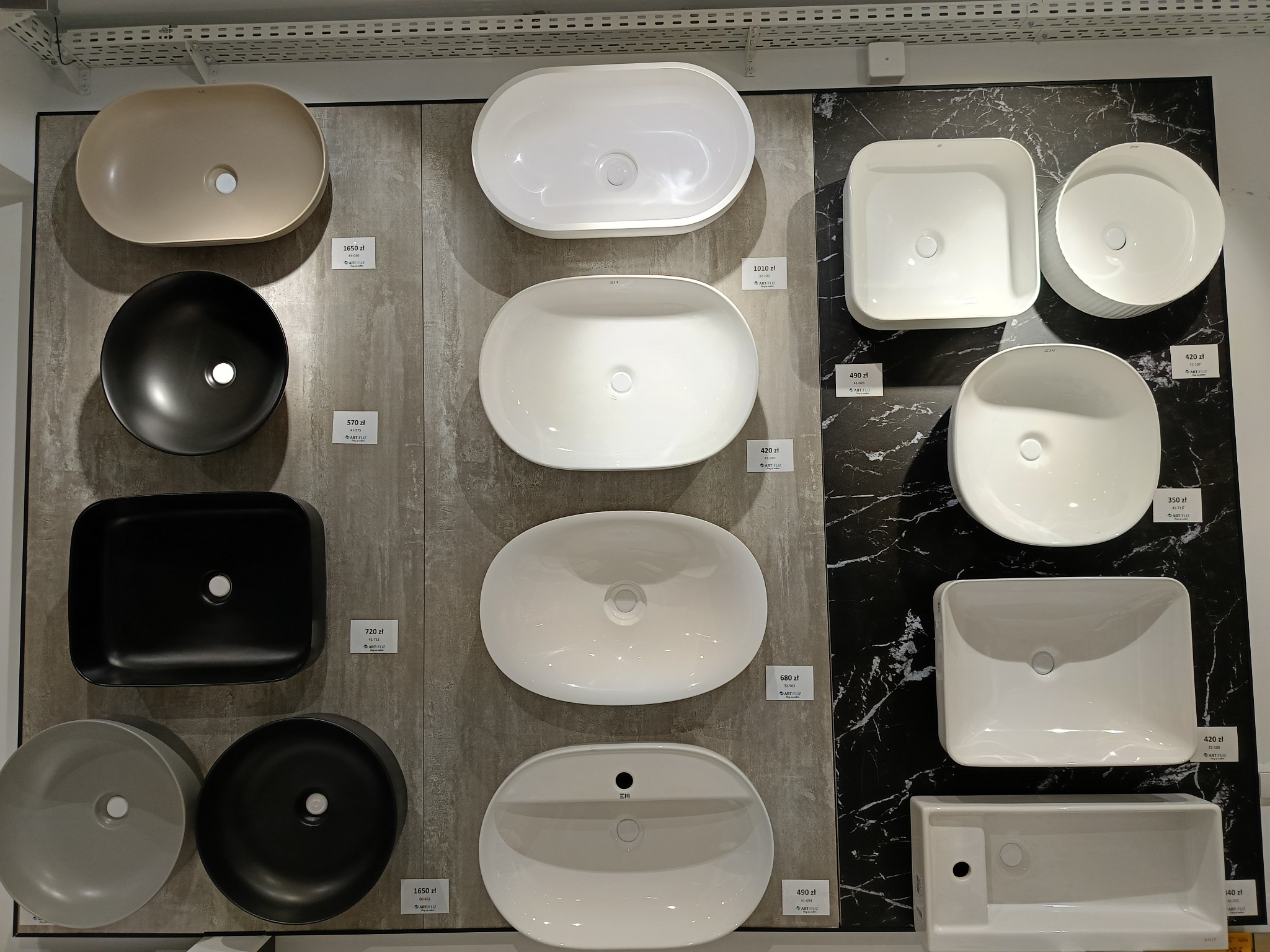Ekspozycja umywalek łazienkowych o różnych kształtach i kolorach, w tym owalnych, okrągłych, kwadratowych i prostokątnych, prezentowanych na ścianach z różnym wykończeniem, wraz z cenami.