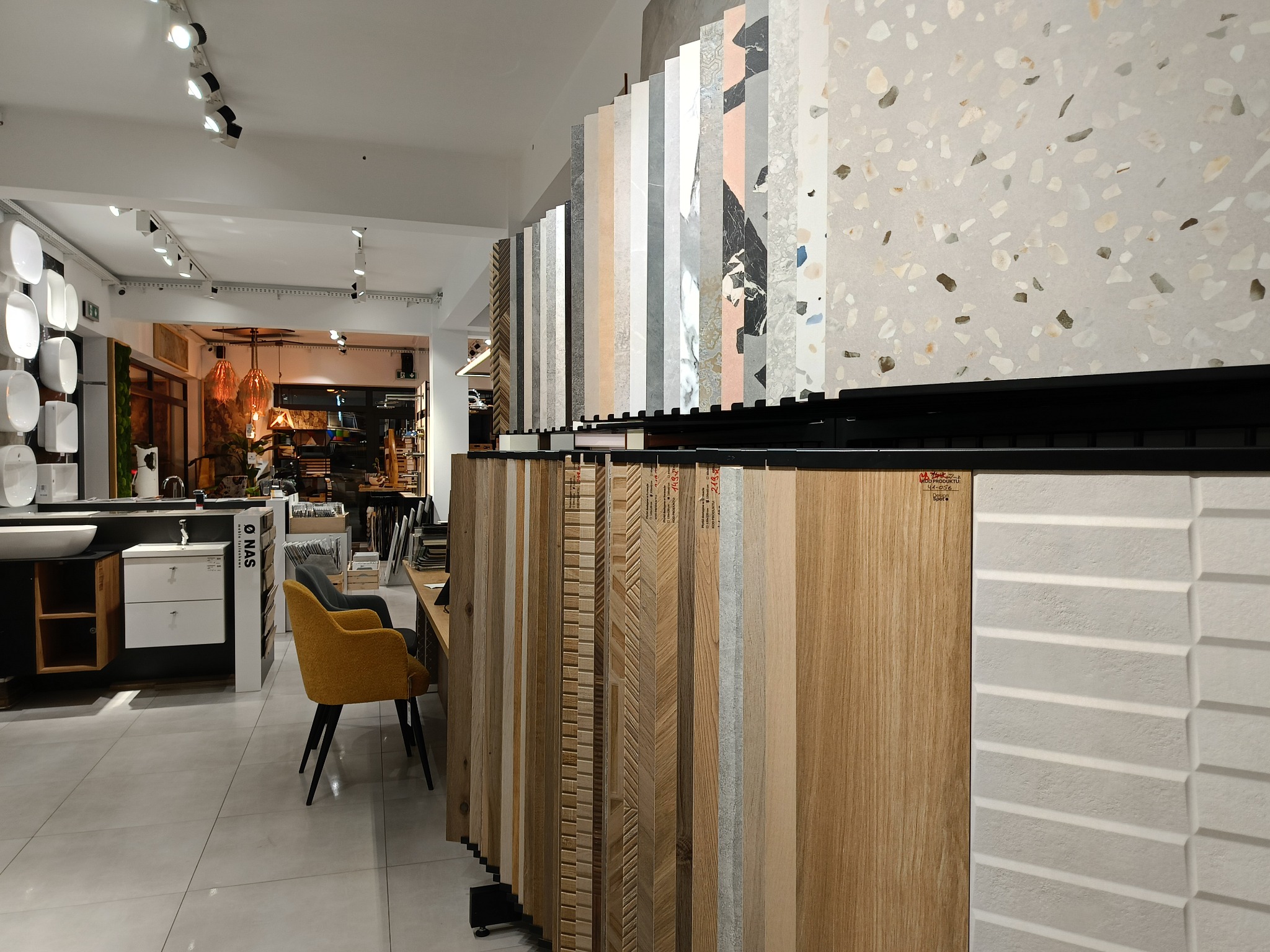 Ekspozycja różnorodnych płytek ceramicznych i paneli podłogowych w salonie, widoczne umywalki i krzesła w tle