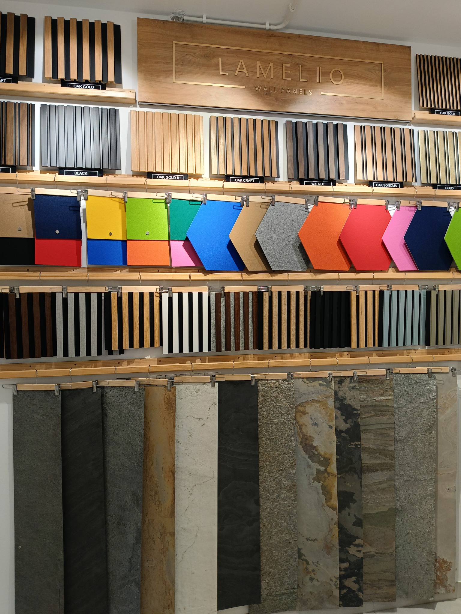 Ekspozycja próbek paneli ściennych Lamelio: drewniane lamele w różnych odcieniach dębu, panele filcowe w kształcie strzałek w żywych kolorach oraz panele kamienne z różnymi fakturami.