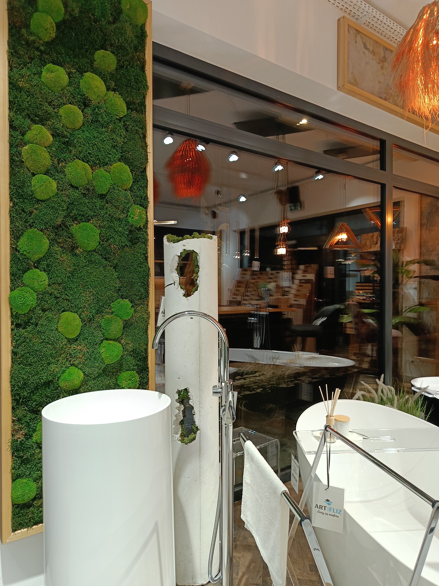 Nowoczesna łazienka z wolnostojącą umywalką cylindryczną, obok panel z mchu, w tle wanna i lampa wisząca.