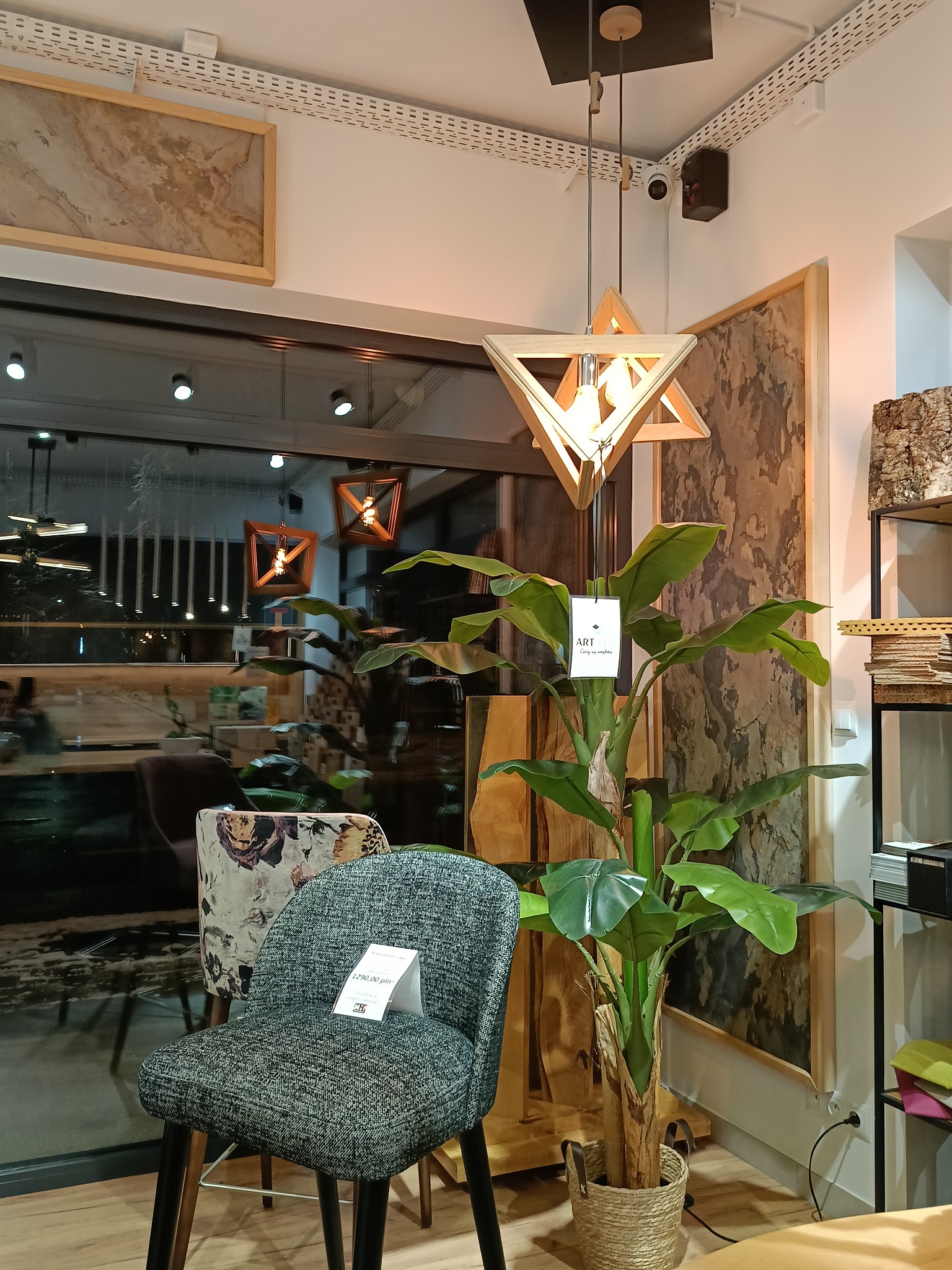Wnętrze sklepu meblowego w Nowym Targu: krzesła z etykietami cenowymi, sztuczna roślina w plecionym koszu, drewniana lampa wisząca o geometrycznym kształcie i dekoracyjny panel ścienny.