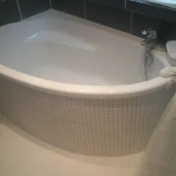 Narożna wanna obudowana mozaiką z drobnych, jasnych płytek, bateria z prysznicem, obok pralka, ciemne płytki na ścianie w tle.