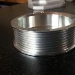 Metalowy, okrągły element z rowkami, prawdopodobnie wykonany metodą CNC, leżący na ciemnym blacie. Widoczne ślady obróbki na powierzchni.