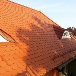 Czerwona dachówka karpiówka na dachu kamienicy z oknami typu wole oko, widok z góry na tle błękitnego nieba.