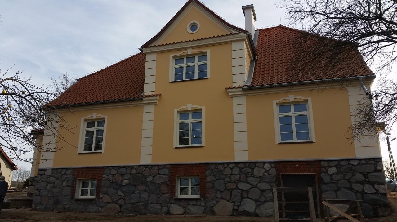 Odrestaurowany dwukondygnacyjny budynek z żółtą elewacją, czerwoną dachówką i kamienną podmurówką, widoczne detale architektoniczne i okna z białymi ramami.