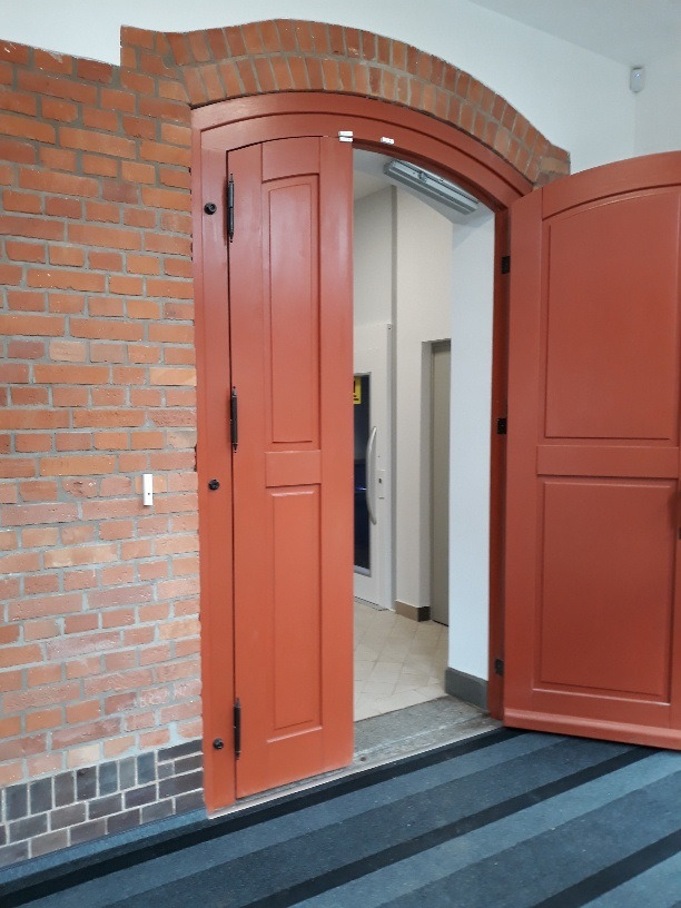 Otwarta, pomarańczowa, dwuskrzydłowa brama łukowa z ceglanym obramowaniem, prowadząca do jasnego wnętrza z windą.