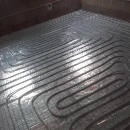 Ułożone rury systemu ogrzewania podłogowego na folii z siatką, widoczne ściany z cegły.