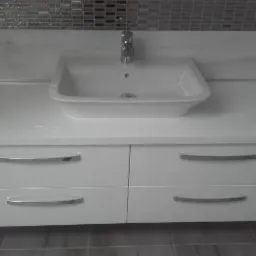 Biała umywalka nablatowa z chromowaną baterią na szafce z czterema szufladami, tło stanowi mozaika ścienna w odcieniach szarości.