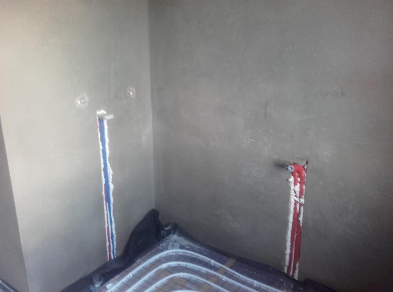 Montaż rur ogrzewania podłogowego na czarnej folii izolacyjnej, widoczne pionowe rury w ścianie z wyciętym tynkiem i śladami po instalacji.