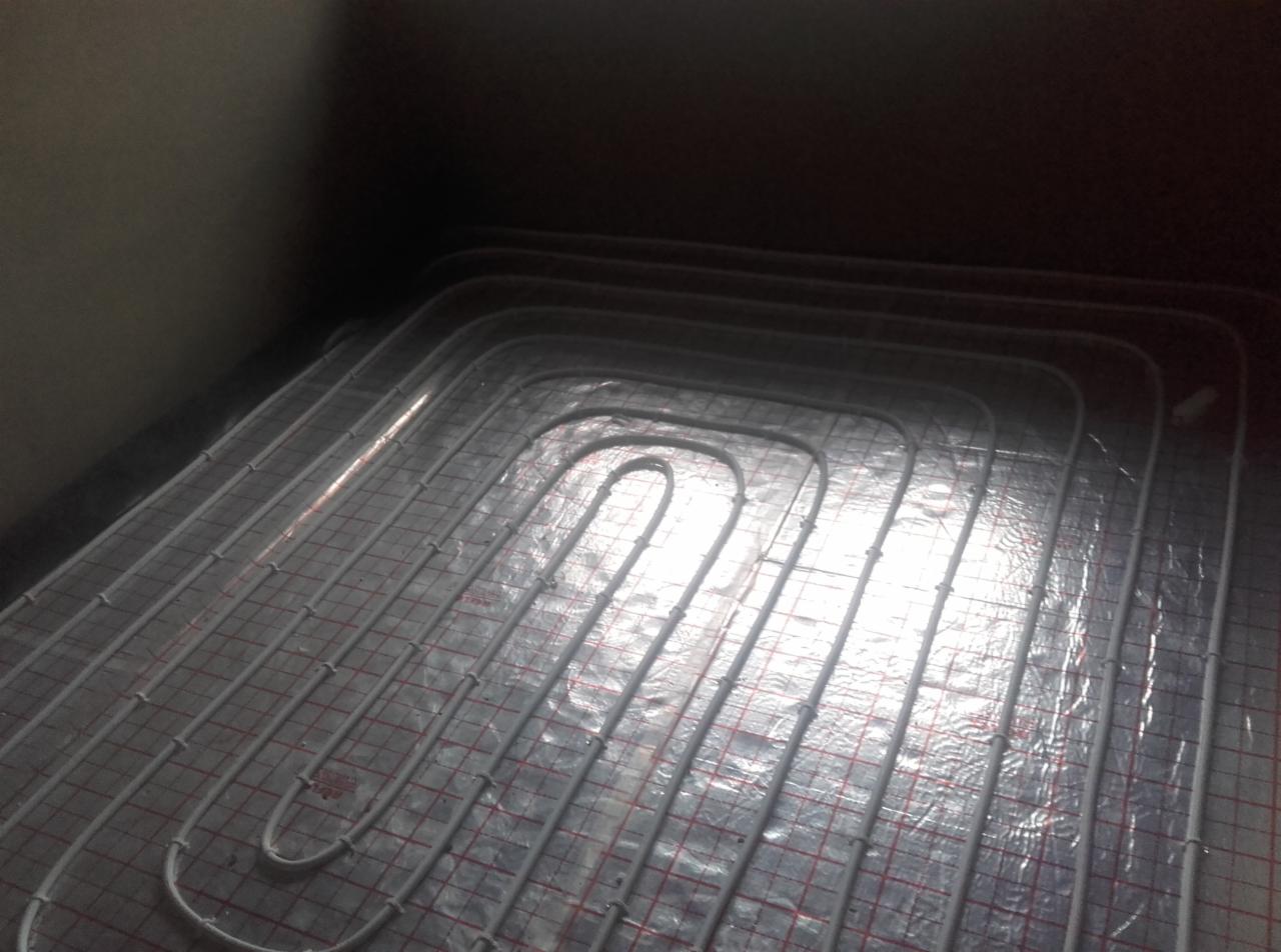 Białe rury układane w pętlach na srebrnej folii z czerwoną siatką, instalacja podłogowa w ciemnym pomieszczeniu przed zalaniem wylewką.