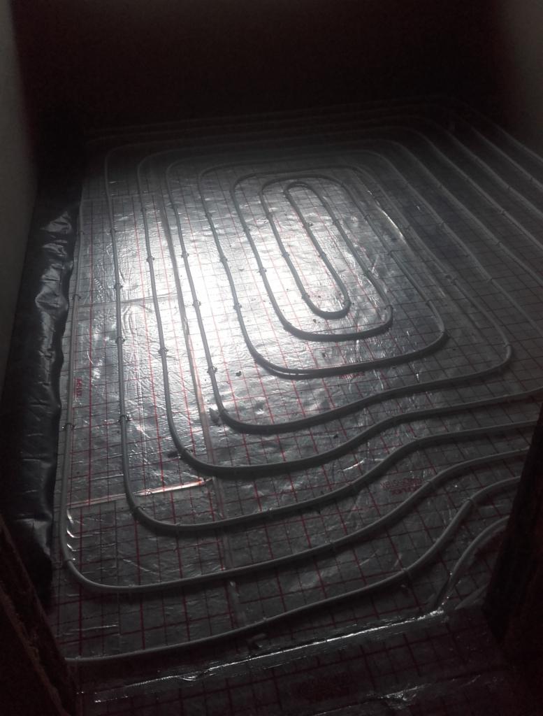 Montaż ogrzewania podłogowego: rury grzewcze ułożone na folii z siatką w ciemnym pomieszczeniu, widoczne ślady wilgoci.