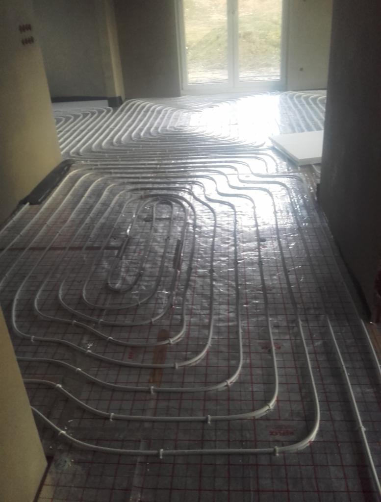 Białe rury systemu ogrzewania podłogowego ułożone na siatce w surowym wnętrzu, widok na okno i niezabudowany teren.