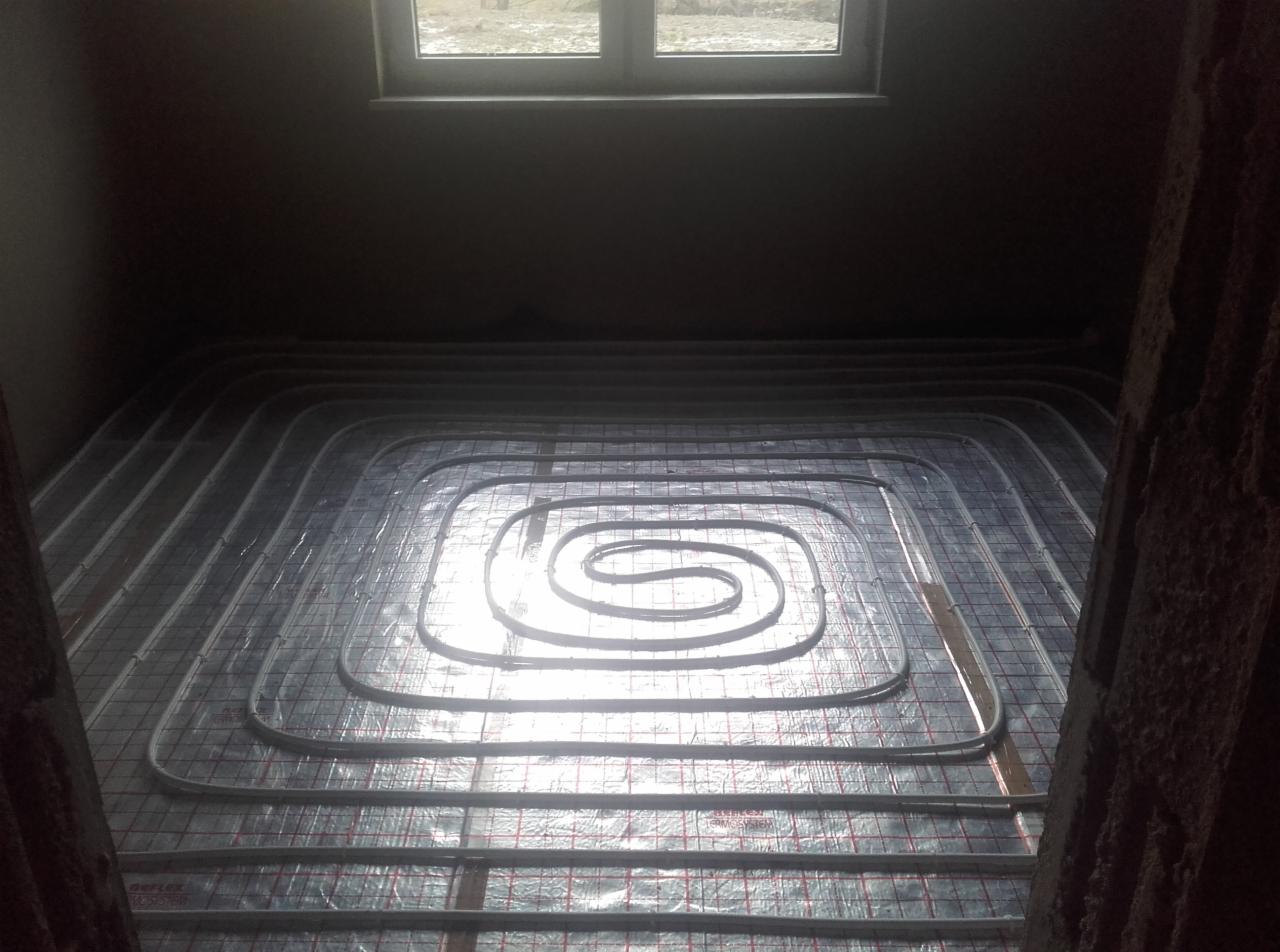 Rozłożone rury ogrzewania podłogowego na srebrnej folii z czerwoną siatką, widoczne okno w tle, surowy stan pomieszczenia.