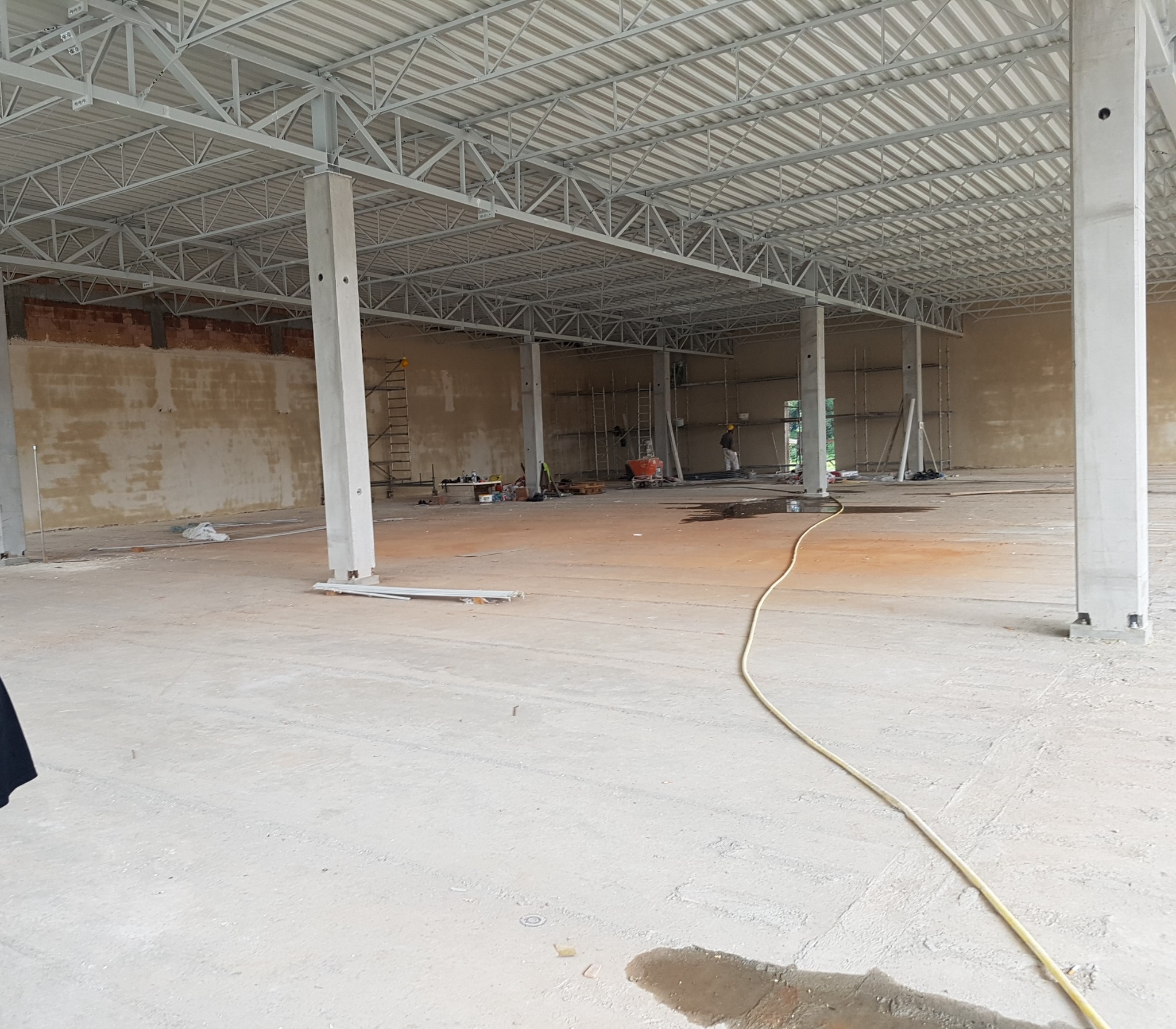 Wnętrze hali w trakcie budowy: surowy beton, metalowa konstrukcja dachu, widoczne słupy nośne, fragmenty ścian z cegły i pracownicy na rusztowaniach w tle.