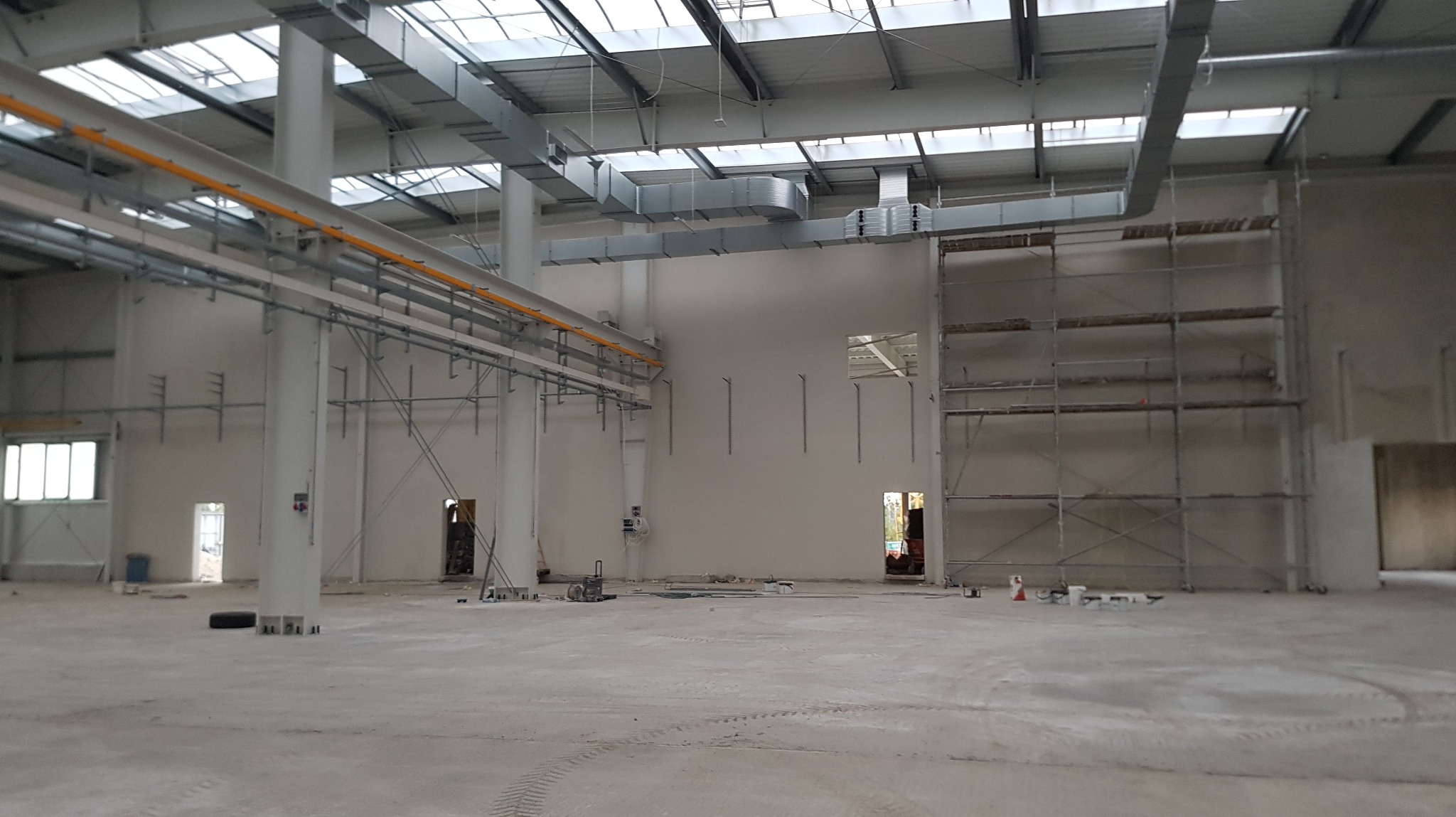 Wnętrze hali przemysłowej w trakcie prac wykończeniowych: widoczne rusztowania, instalacje pod sufitem, surowy beton na podłodze i ściany przygotowane do tynkowania.