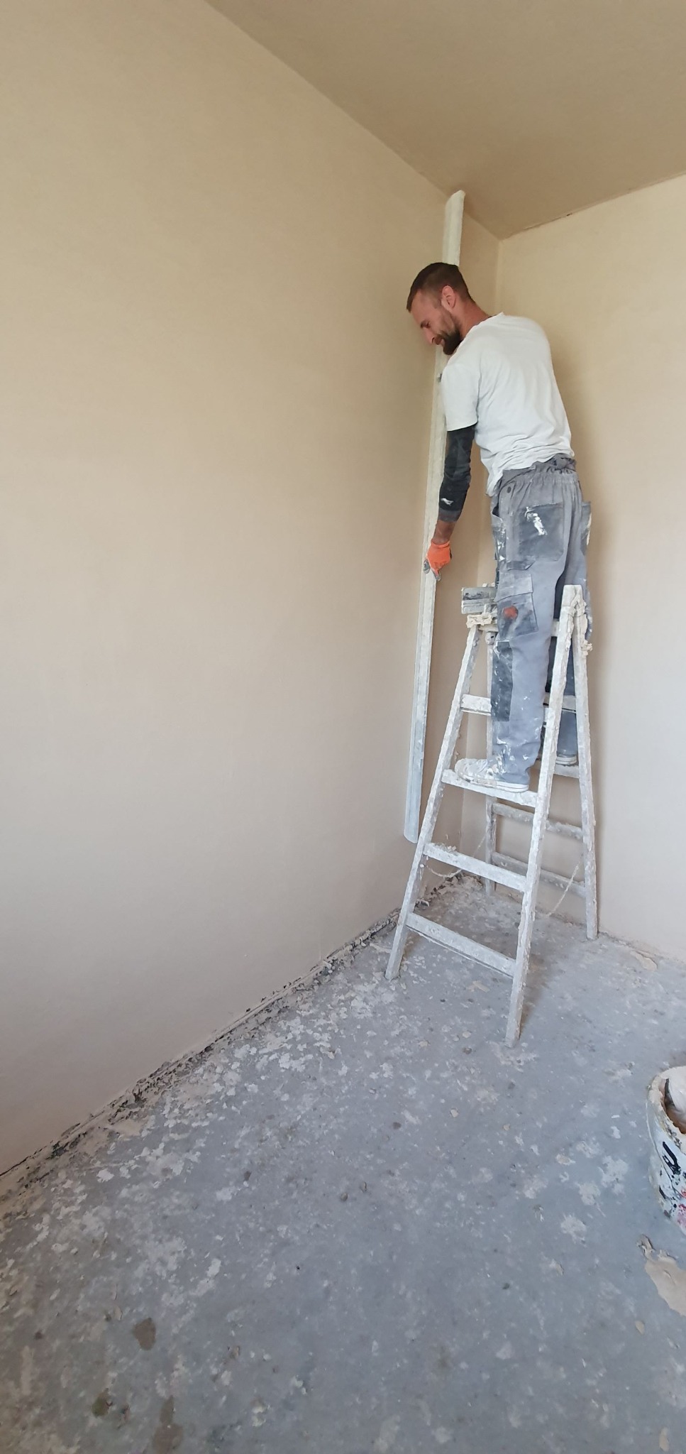 Pracownik na drabinie wyrównuje ścianę po nałożeniu tynku. Widoczne narzędzia i świeżo otynkowana ściana w jasnym odcieniu beżu. Podłoga częściowo zabrudzona.