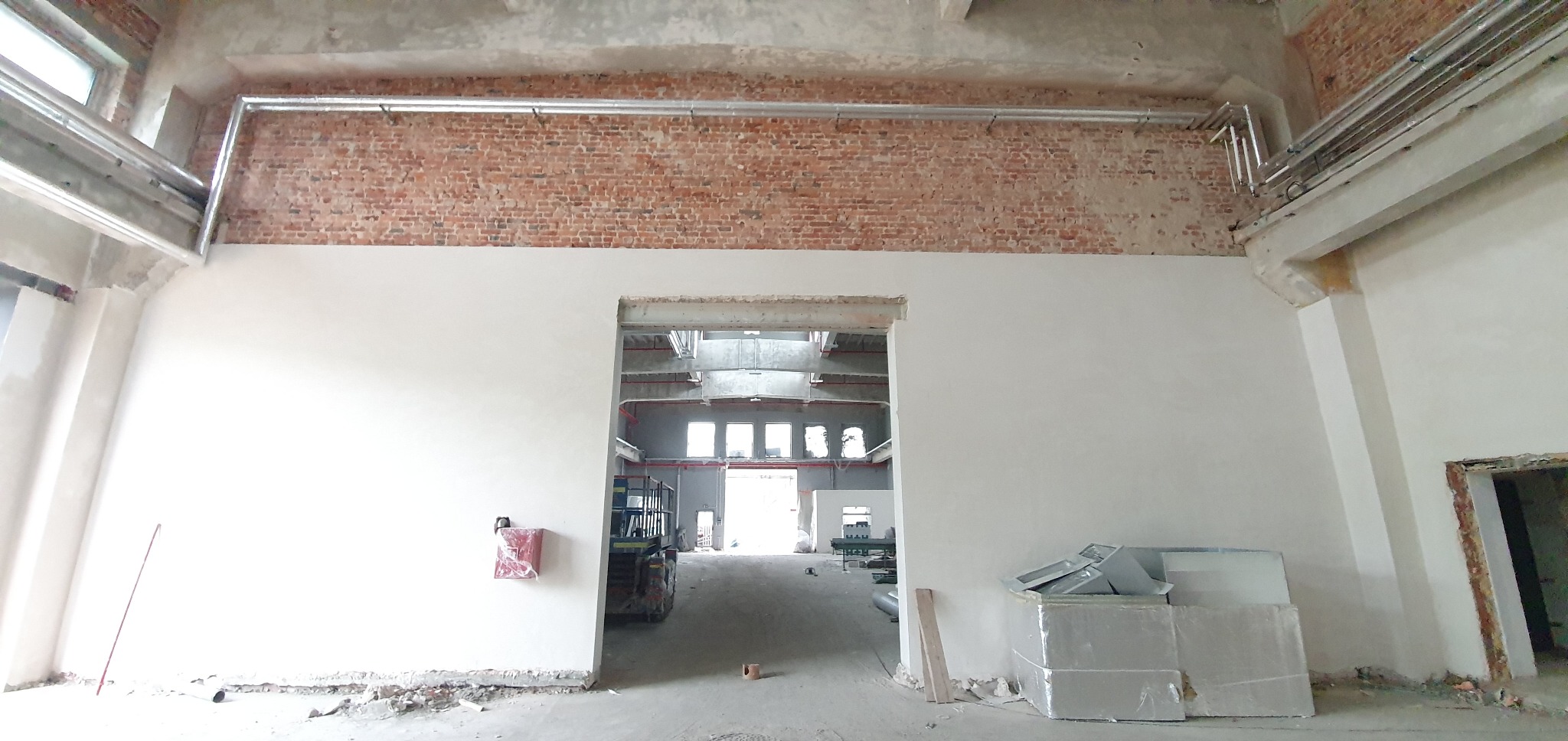 Wnętrze hali po tynkowaniu maszynowym: gładkie, białe ściany do połowy wysokości, powyżej odsłonięta cegła. Widoczne instalacje pod sufitem i materiały budowlane.