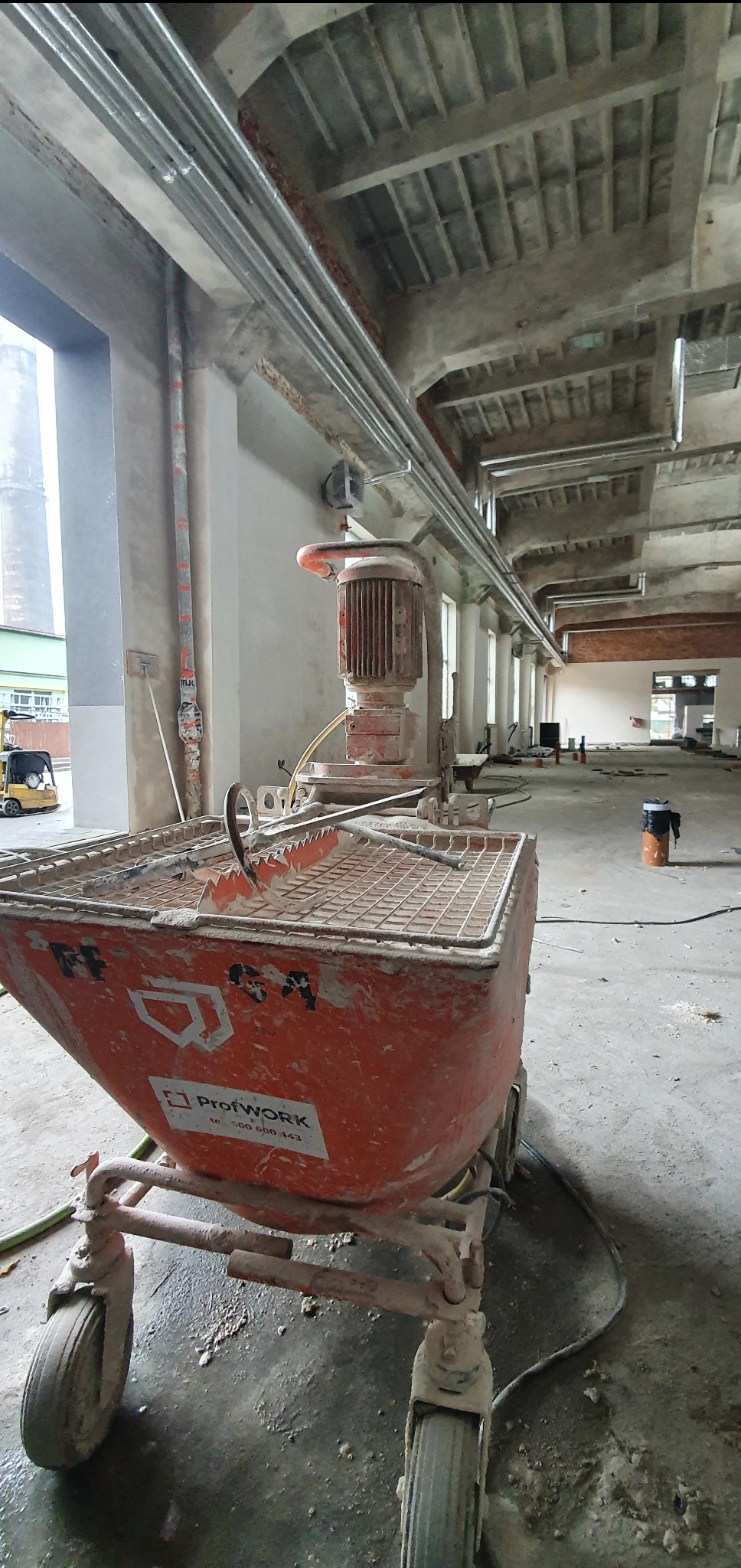 Pomarańczowa maszyna do tynków maszynowych ProfWORK na terenie budowy, widoczne rury pod sufitem i surowy stan wykończenia wnętrza. W tle widoczny wózek widłowy.