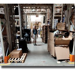 AVO technika sp z o o - Pracownik w magazynie, wyposażony w urządzenie pomiarowe, stoi na tle regałów z towarem. Widoczne palety i kartony. Oświetlenie magazynowe.