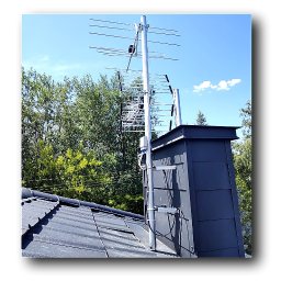 AVO technika sp z o o - Antena telewizyjna zamontowana na kominie, widok z dołu na tle błękitnego nieba i zieleni drzew. Instalacja antenowa na dachu z ciemnej dachówki.