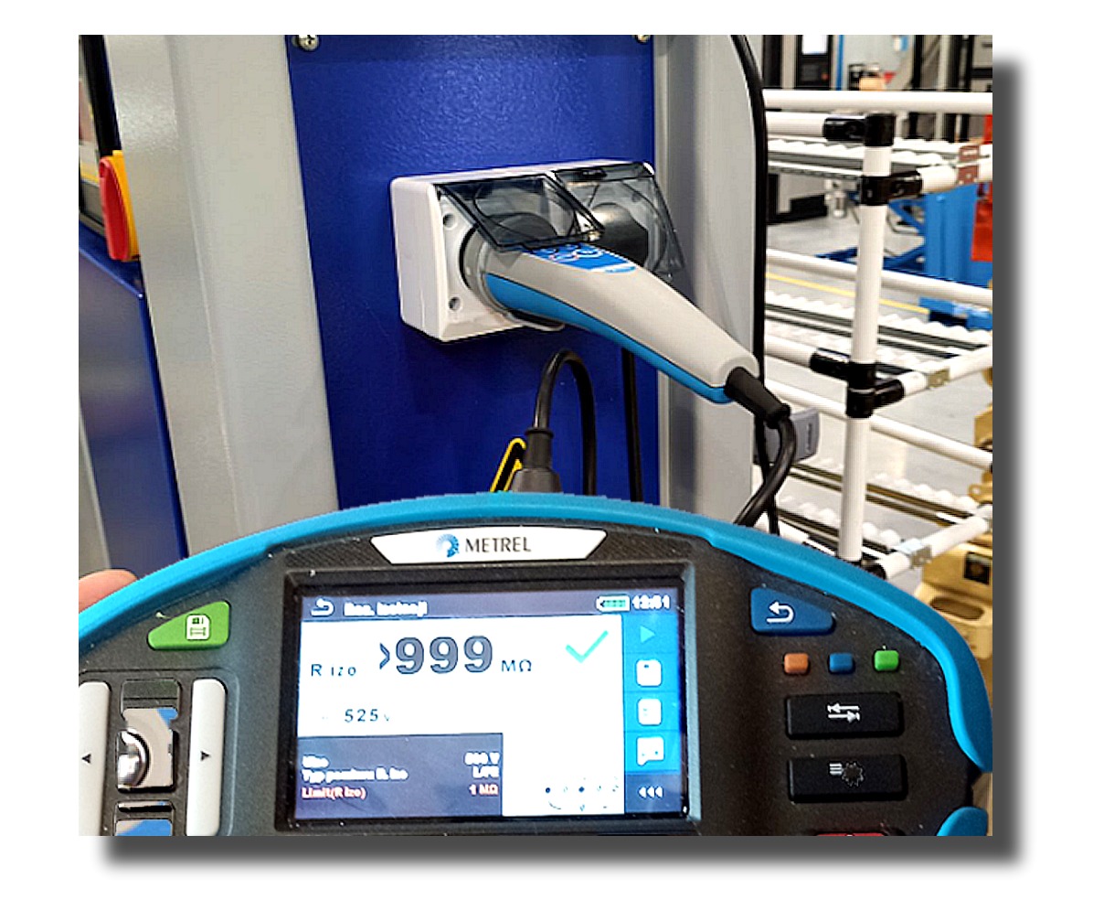 Miernik Metrel wskazuje wynik pomiaru rezystancji izolacji gniazda elektrycznego. Widoczny wynik powyżej 999 MΩ. Tło stanowią elementy instalacji elektrycznej.