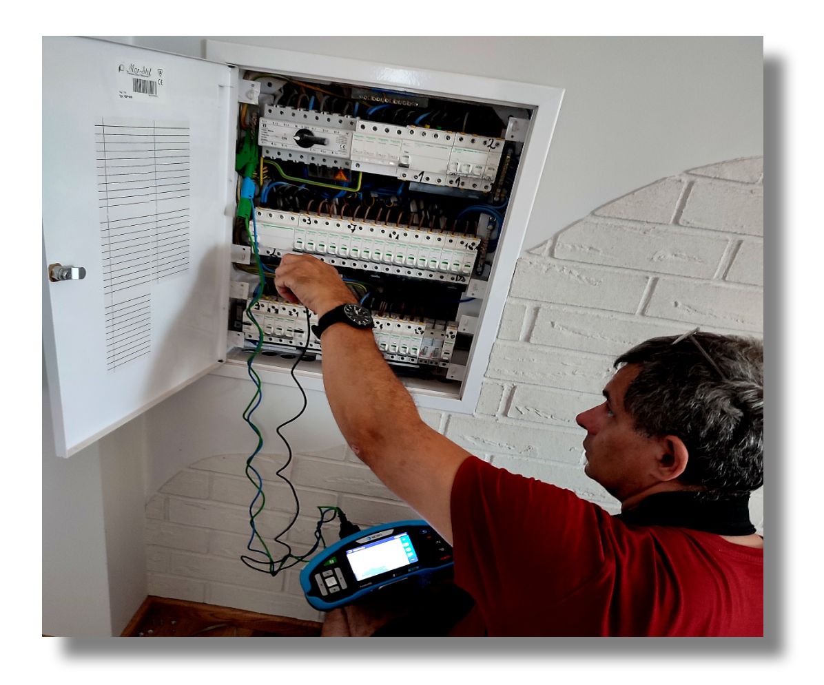 Elektryk w trakcie pomiarów w rozdzielnicy elektrycznej, używając specjalistycznego miernika. Widoczne przewody, elementy instalacji i biała ściana z cegieł.