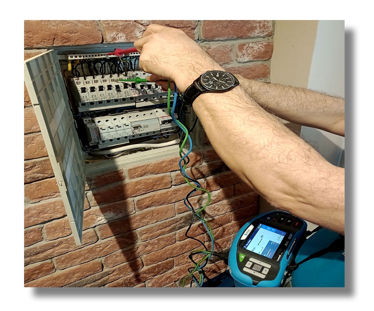 Elektryk mierzy parametry w rozdzielnicy elektrycznej na tle ceglanej ściany. Widoczne przewody, miernik i zegarek na ręce.