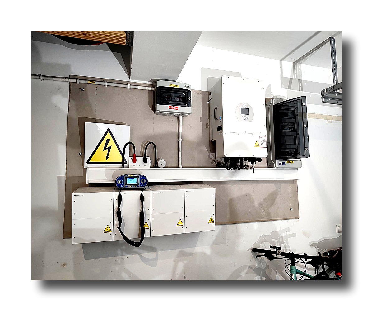 Instalacja fotowoltaiczna w garażu: falownik, rozdzielnica, akumulatory z symbolem ostrzegawczym i miernik elektryczny na tle ściany. Widoczny rower.