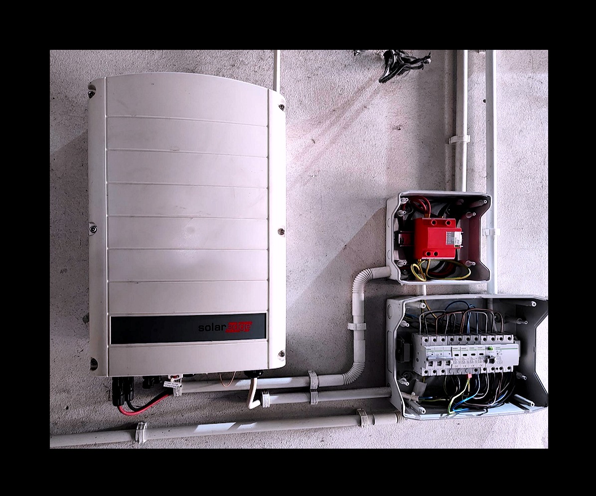 Biały falownik SolarEdge na szarej ścianie, z widocznymi przewodami i skrzynkami elektrycznymi. Instalacja fotowoltaiczna wewnątrz budynku.