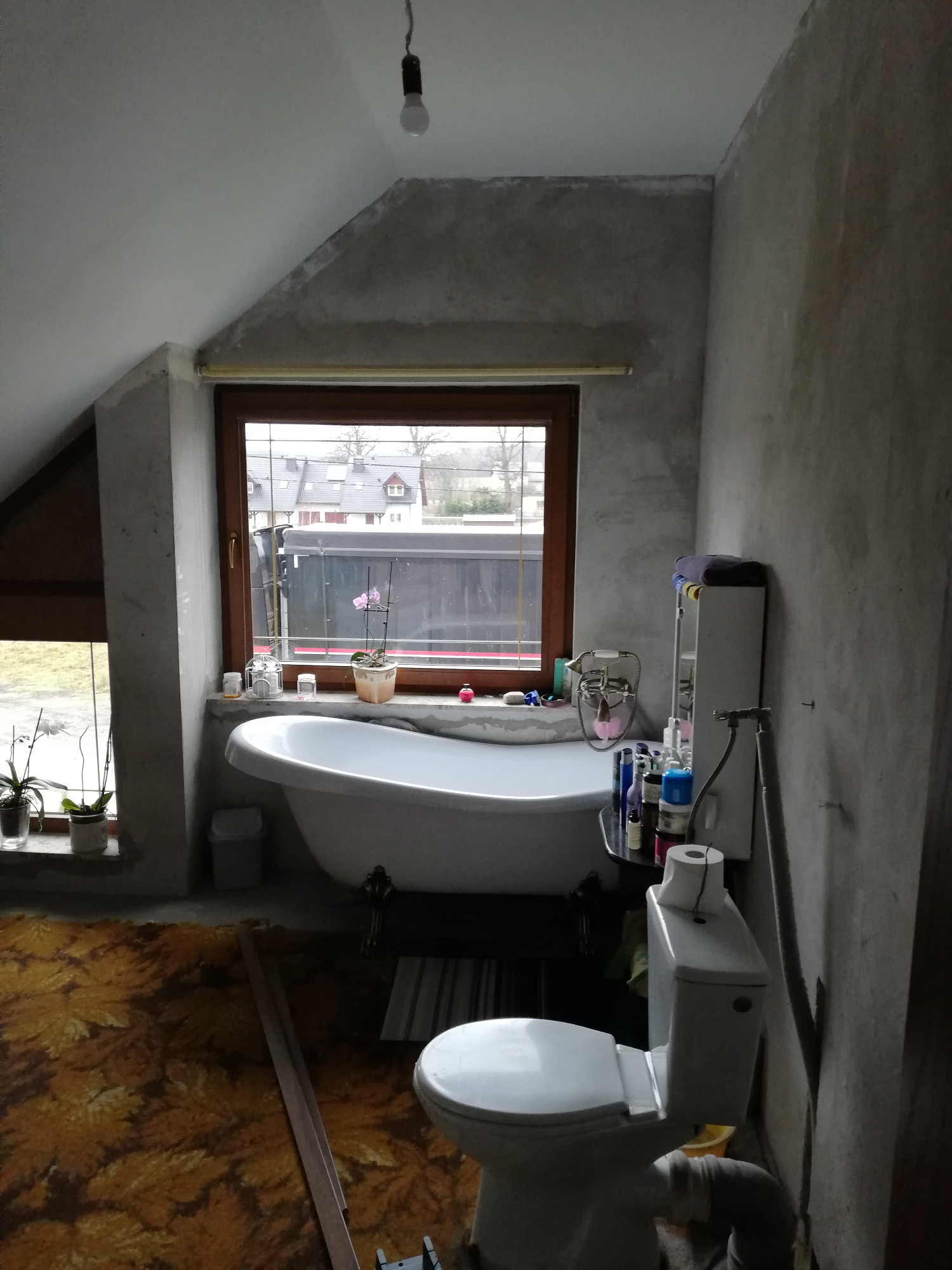 Łazienka na poddaszu w trakcie remontu: ściany po szpachlowaniu, wolnostojąca wanna na ozdobnych nóżkach, toaleta, okna z widokiem na zabudowę miejską, tymczasowe oświetlenie.