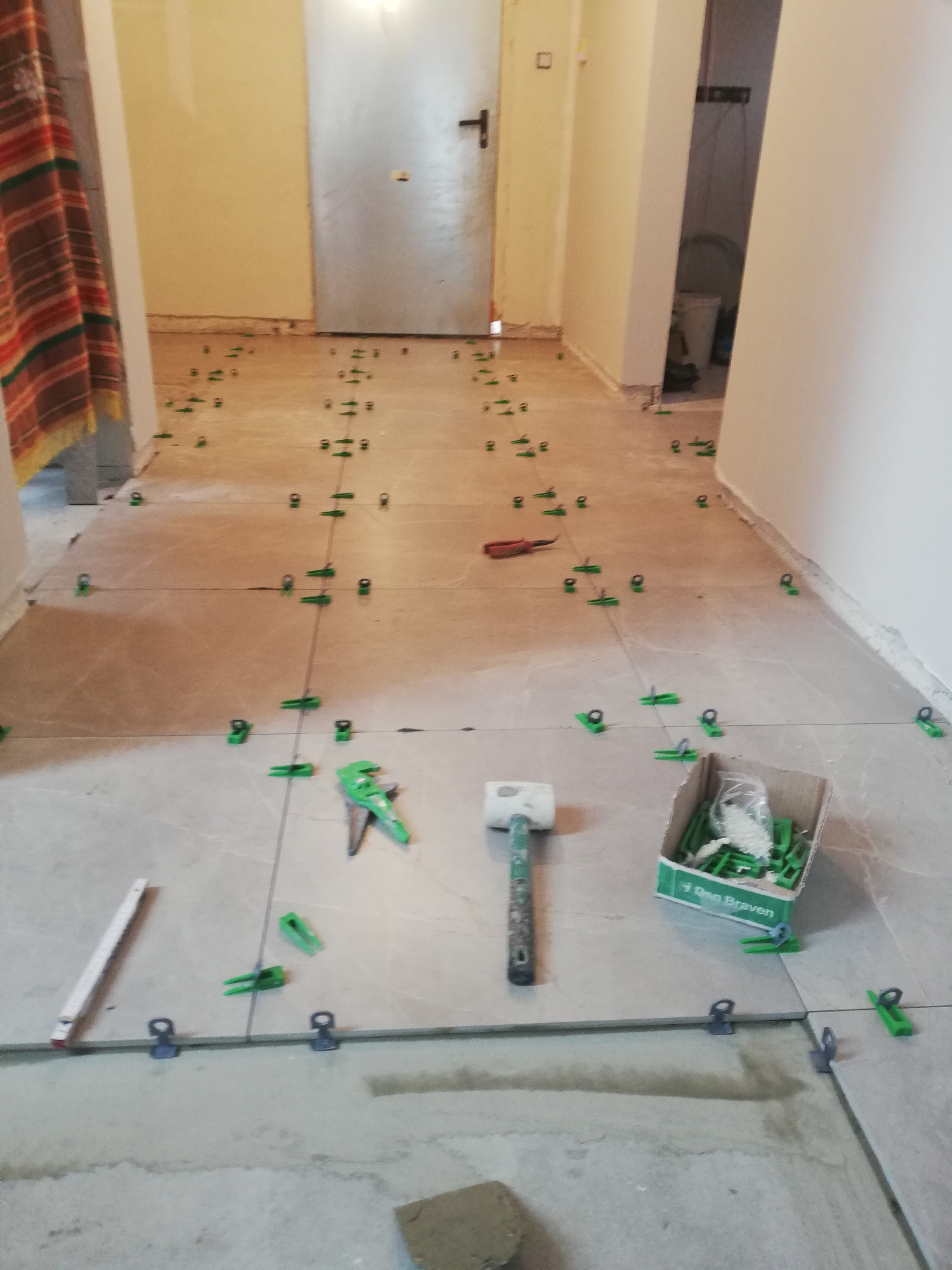 Układanie płytek podłogowych w korytarzu, widoczne narzędzia: młotek, poziomica, pistolet do klinów, kliny poziomujące i sznurki wyznaczające linie, płytki w kolorze beżowym.