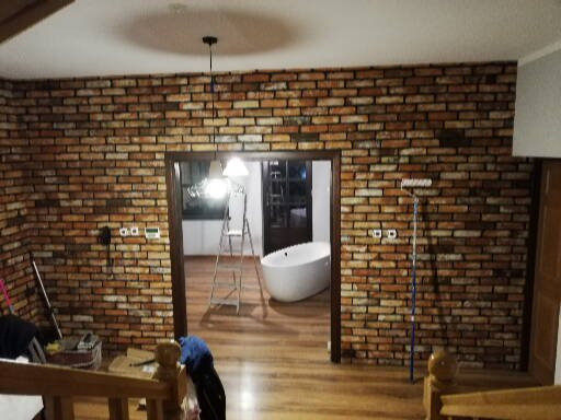 Ściana wyłożona cegłą klinkierową w łazience, widoczna wanna wolnostojąca i drabina malarska, oświetlenie sufitowe, perspektywa z dołu schodów.