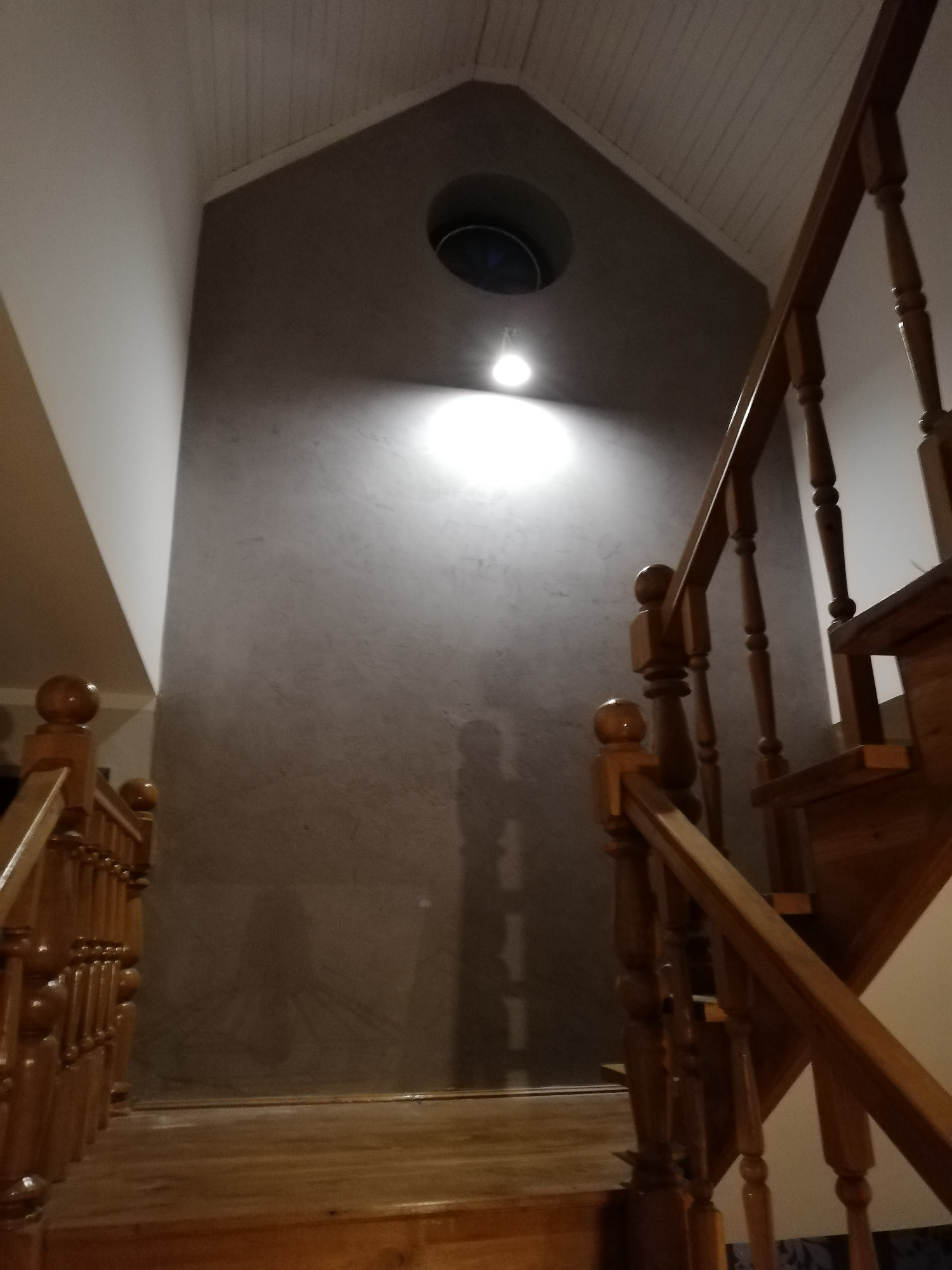 Klatka schodowa z drewnianymi schodami i szarą ścianą z widoczną fakturą, oświetlona punktowym światłem z sufitu, z okrągłym otworem wentylacyjnym w górnej części ściany.