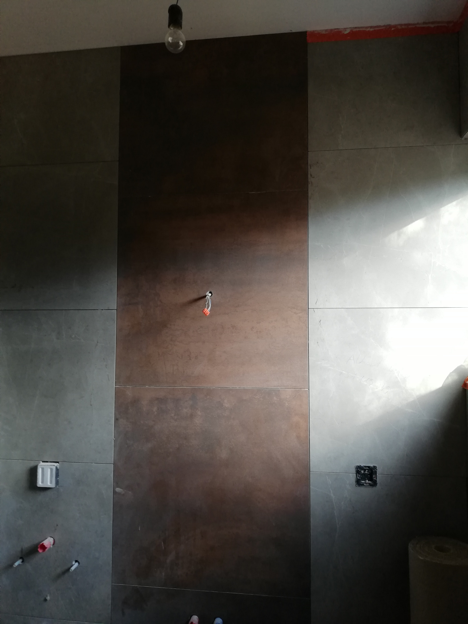 Ściana wyłożona płytkami w dwóch odcieniach szarości i brązu, widoczne przygotowane punkty instalacyjne, surowa żarówka zwisająca z sufitu, fragmenty taśmy malarskiej na górze.