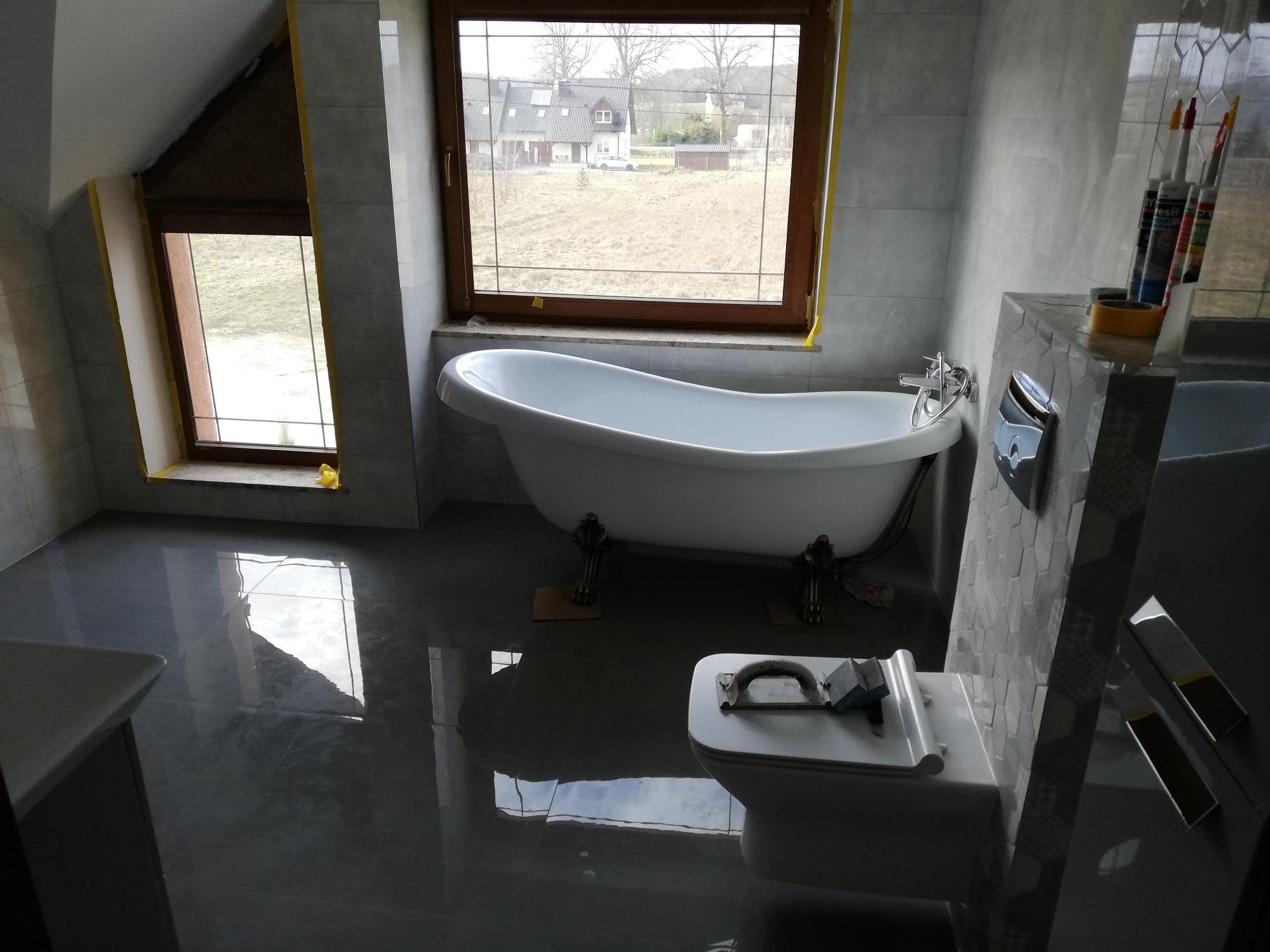 Nowoczesna łazienka na poddaszu z wolnostojącą wanną na ozdobnych nóżkach, umywalką, toaletą z narzędziami na desce, wykończona szarą płytką, z oknem z widokiem na krajobraz, zabezpieczona taśmą...