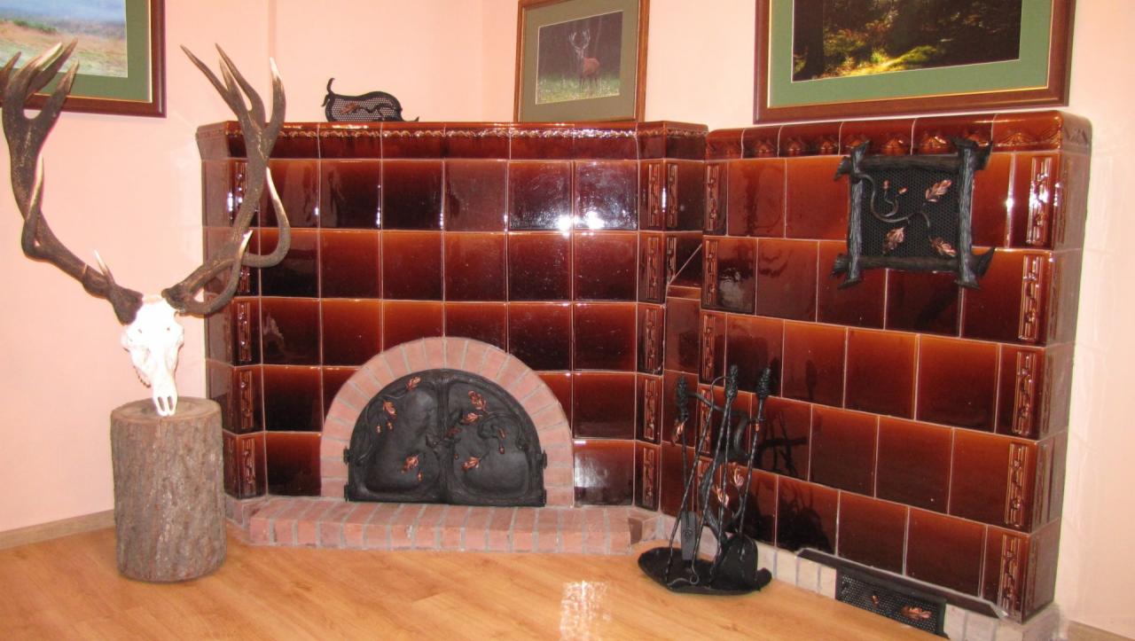 Rustykalny piec kaflowy z dekoracyjną kratką i akcesoriami kominkowymi, uzupełniony trofeum myśliwskim na tle drewnianej podłogi i jasnych ścian.