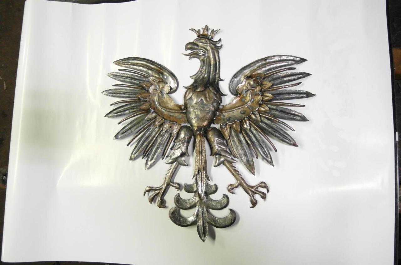 Metalowy, srebrno-złoty orzeł w koronie, z rozpostartymi skrzydłami, umieszczony na białym tle. Detal kowalstwa artystycznego.