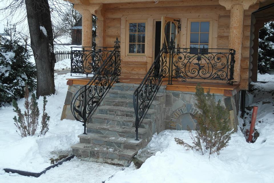 Zimowy widok na wejście do drewnianego domu z kamiennymi schodami i ozdobnymi, kutymi balustradami w kolorze czarnym.